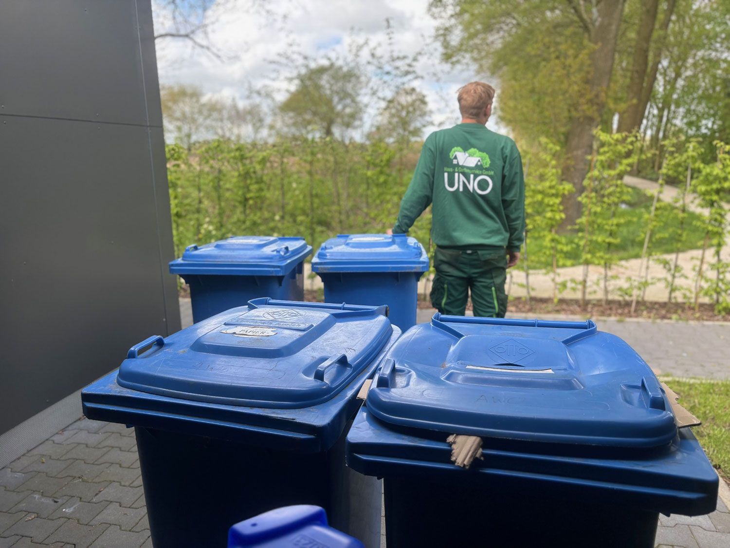 Drei blaue Mülltonnen und eine weitere Mülltonne  wird von einem Mitarbeiter gezogen